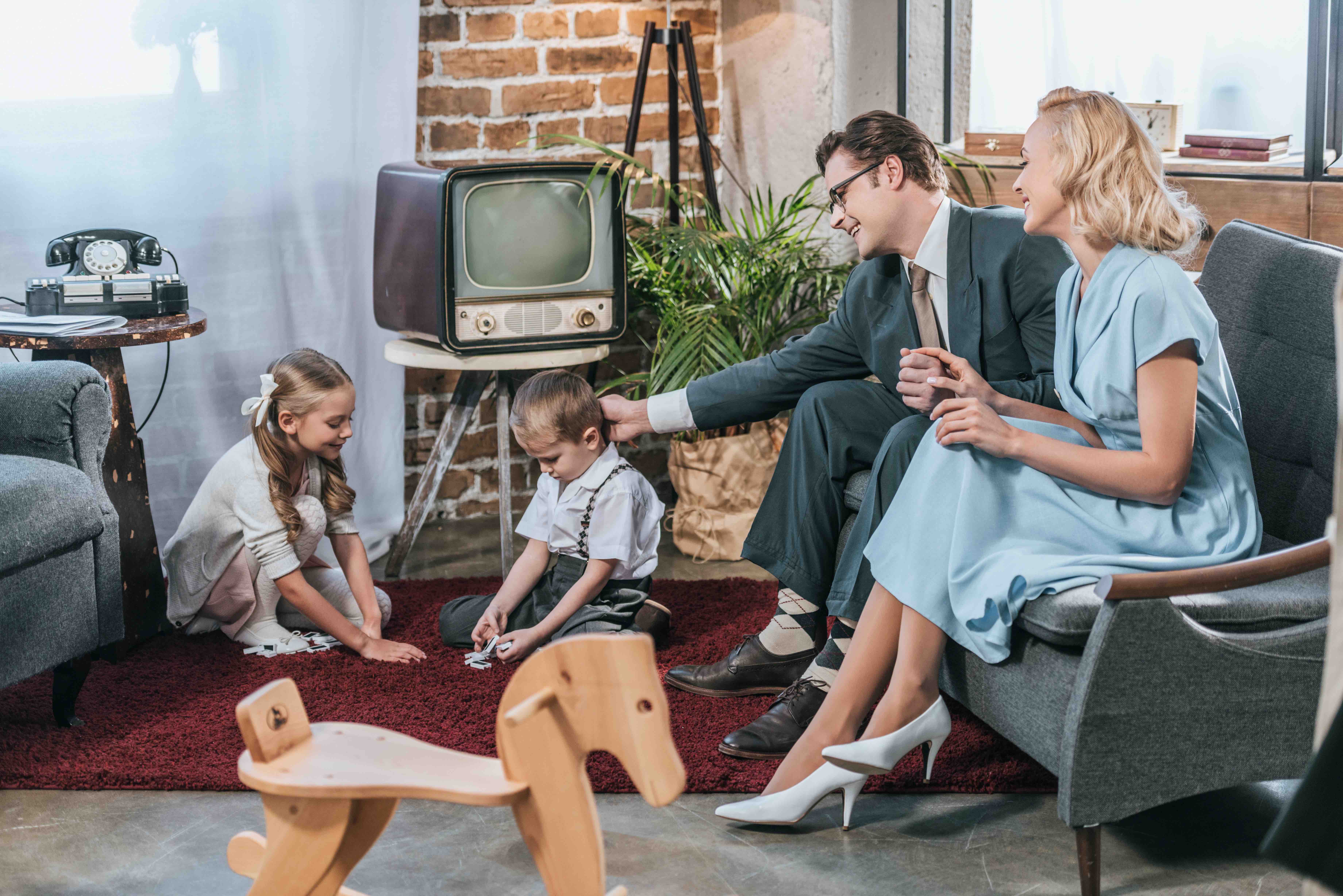 glückliche Eltern sitzen auf dem Sofa und schauen sich kleine Kinder an, die zu Hause mit Dominosteinen spielen, Stil der 50er Jahre glückliche Eltern sitzen auf dem Sofa und schauen sich kleine Kinder an, die zu Hause mit Dominosteinen spielen, Stil der 50er Jahre