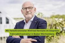 Das Bild zeigt eine Person mit Brille und einem Anstecker, die direkt in die Kamera blickt. Über der Person steht in einer Schriftleiste: "Mit Verkehrsminister Winfried Hermann im Gespräch: Fahrradinfrastruktur in Baden-Württemberg". Im Hintergrund ist unscharf eine Gebäudestruktur zu erkennen.