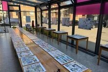 Ein heller Ausstellungsraum mit großen Glasfronten zeigt zahlreiche Comics auf langen Tischen und einzelnen Ausstellungstischen. An den Wänden hängen weitere Exponate. Im Hintergrund sieht man Gebäude und Fahrzeuge draußen.