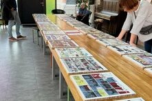 Zwei Besucher*innen betrachten aufmerksam Comics, die auf langen Tischen in einem lichtdurchfluteten Raum ausgelegt sind. Eine weitere Person blättert durch ein Comic-Heft.