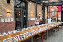 In einem hellen Ausstellungsraum betrachten Besucher*innen Comics, die auf einem langen Tisch ausgestellt sind. Die Wände bestehen aus Backsteinen, und große Türen und Fenster verbinden Innen- und Außenraum sichtbar miteinander.