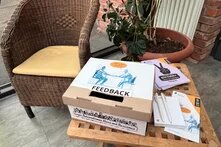 Ein kleiner Tisch mit einer Feedback-Box aus Pappe, beschriftet mit 'Gegen Antisemitismus, Hass und Rassismus'. Daneben liegen Broschüren und Schreibmaterial. Im Hintergrund ein Korbsessel und eine Pflanze.