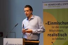 Ein Mann in einem hellblauen Hemd steht an einem Pult und hält eine Rede. Das Pult trägt das Logo der Akademie der Diözese Rottenburg-Stuttgart. Im Hintergrund ist ein Banner der Heinrich Böll Stiftung mit dem Zitat: „Einmischung ist die einzige Möglichkeit, realistisch zu bleiben.“