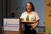 Eine Frau in einem weißen, kurzärmeligen Oberteil mit Brille steht an einem Pult und spricht. Das Pult trägt das Logo der Akademie der Diözese Rottenburg-Stuttgart. Im Hintergrund ist ein Banner der Heinrich Böll Stiftung mit dem Zitat: „Einmischung ist die einzige Möglichkeit, realistisch zu bleiben.“