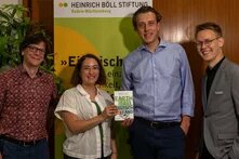Vier Personen stehen vor einem Banner der Heinrich Böll Stiftung. Die Frau in der Mitte hält ein Buch mit dem Titel „Earth for All Deutschland: Aufbruch in eine Zukunft für Alle“. Das Banner im Hintergrund zeigt das Zitat: „Einmischung ist die einzige Möglichkeit, realistisch zu bleiben.“