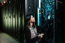 Eine Person in einem Rechenzentrum bedient einen offenen Serverschrank mit zahlreichen Kabeln. Die Person trägt eine gemusterte Weste über einem dunklen Oberteil und hält ein Tablet oder ein Werkzeug in der Hand. Die Umgebung ist dunkel, und die Serverracks sind beleuchtet. Die Szene vermittelt den Eindruck von Wartungs- oder Installationsarbeiten an IT-Infrastruktur.