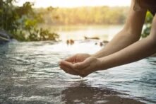 Zwei Hände, die über einem flachen Wasserbecken gehalten werden, aus dem Wasser tropft. Im Hintergrund ist ein See mit Bäumen und Vögeln zu sehen.