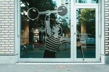 Silhouette eines muskulösen Armes im gestreiften Sportdress, der eine Hantelscheibe mit zwei Verkehrsschildern – ein Rundverkehrszeichen und ein „Vorfahrt gewähren“-Schild – nach oben stemmt. Die Folie ist auf einer Glastür angebracht, im Hintergrund sind Straßen, Bäume und Gebäude zu erkennen. 