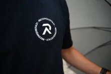 Rundes Logo auf schwarzem T-Shirt mit weißer Schrift und Grafik: Großes „R“ in der Mitte, umgeben vom kreisförmigen Text „RECONQUISTA21 - RECONQUISTA21“