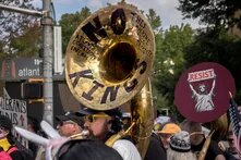 Claude hat geantwortet: Protestfoto einer Demonstration in Atlanta.Protestfoto einer Demonstration in Atlanta. Im Vordergrund eine Person, die eine Tuba spielt, auf deren Schalltrichter die Aufschriften „NO KINGS", „THIS MACHINE KILLS FASCISTS" und „ATLANTA COALITION ENSEMBLE" zu lesen sind. Im Hintergrund Demonstrierende mit Schildern, darunter „RESIST" mit einer Darstellung der Freiheitsstatue, „MIGRANTS […] US" sowie US-amerikanische Flaggen.