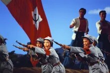 Szenenfoto einer chinesischen Revolutionsoper. Mehrere Tänzerinnen in grauen Militäruniformen mit roten Armbinden und Mützen mit rotem Stern zielen mit Gewehren nach vorne, mit expressiven Gesichtsausdrücken. Im Hintergrund eine rote Fahne mit Hammer-und-Sichel-Symbol sowie zwei weitere Darsteller vor blauem Bühnenhimmel.