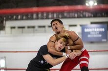 Zwei Ringkämpfer im Clinch auf einem Boxring in Brasilien. Eine Person im schwarzen Shirt hält die andere, die rote Sporthose trägt, in einem Würgegriff. Beide haben die Augen geschlossen und den Mund geöffnet. Im Hintergrund eine Sporthalle mit portugiesischsprachigen Werbebannern.