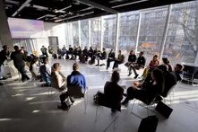 Eine Personengruppe sitzt in einem Stuhlkreis in einem Konferenzraum. Auf einer Leinwand wird eine Präsentation projiziert.