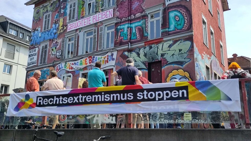 Mehrere Personen stehen vor einem mit Graffiti bemalten Haus mit der Aufschrift „ALL COLORS ARE BEAUTIFUL“, „GEFLÜCHTETE BLEIBEN“ und „KEIN MENSCH IST ILLEGAL“. Im Vordergrund ist ein Banner mit dem Slogan „Rechtsextremismus stoppen“ in schwarzer Schrift auf weißem Grund mit Regenbogenfarben zu sehen.