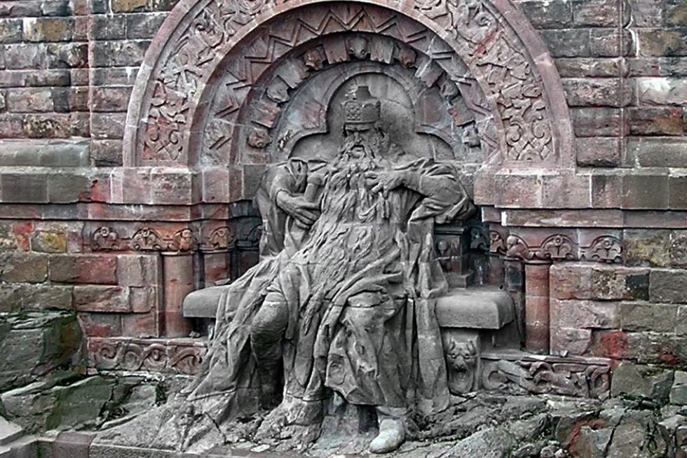 Steinerne Statue eines bärtigen, sitzenden Königs mit Krone vor reich verziertem Hintergrund. Dargestellt ist Kaiser Barbarossa als Teil eines Denkmals.