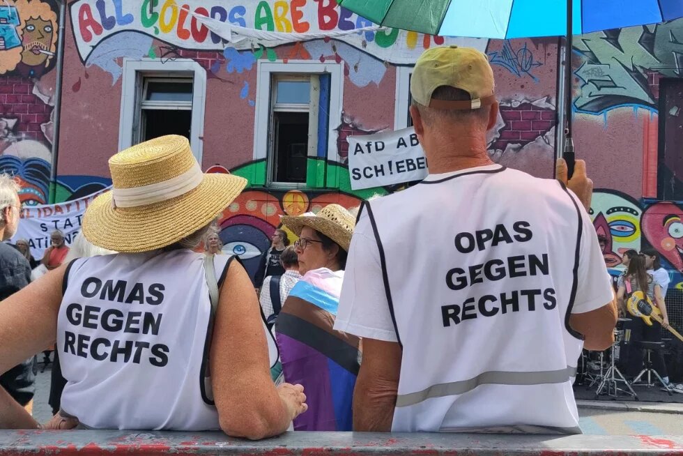 Zwei ältere Personen mit Strohhüten, die Westen mit den Aufschriften „OMAS GEGEN RECHTS“ und „OPAS GEGEN RECHTS“ tragen. Im Hintergrund ein bunt bemaltes Haus mit Graffiti und der Aufschrift „ALL COLORS ARE BEAUTIFUL“. Ein Transparent ruft zur Abschiebung der AfD auf.