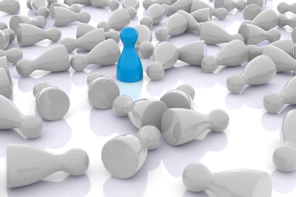 Eine blaue Spielfigur steht aufrecht inmitten zahlreicher umgestürzter, weißer Spielfiguren auf einer glänzenden Oberfläche. Die zentrale Figur hebt sich deutlich durch Farbe und Position ab.