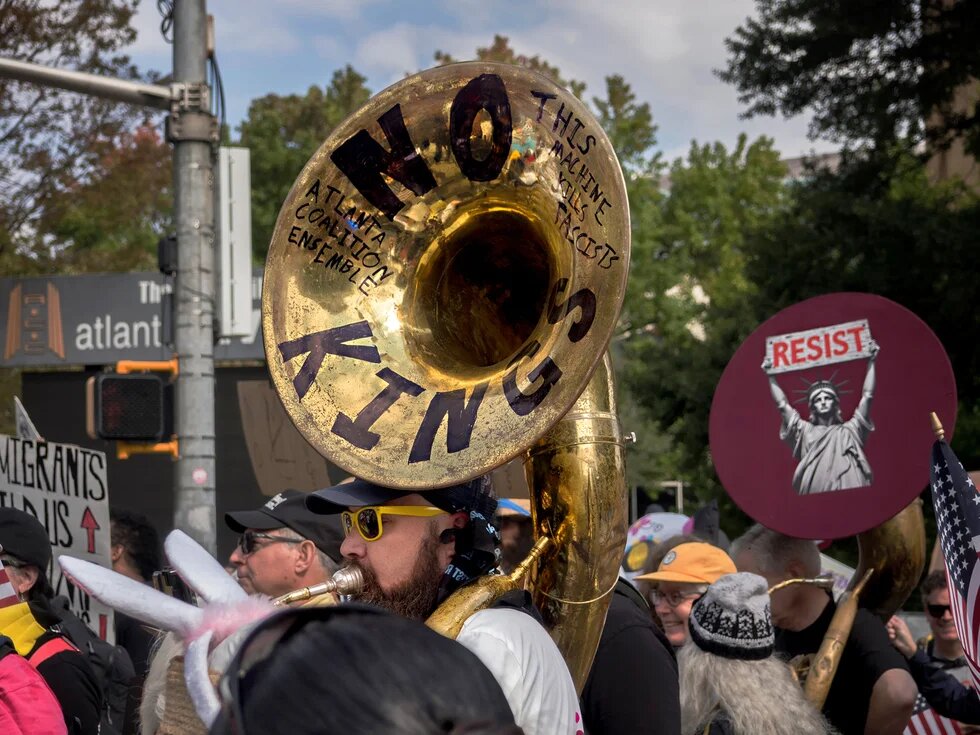 Claude hat geantwortet: Protestfoto einer Demonstration in Atlanta.Protestfoto einer Demonstration in Atlanta. Im Vordergrund eine Person, die eine Tuba spielt, auf deren Schalltrichter die Aufschriften „NO KINGS", „THIS MACHINE KILLS FASCISTS" und „ATLANTA COALITION ENSEMBLE" zu lesen sind. Im Hintergrund Demonstrierende mit Schildern, darunter „RESIST" mit einer Darstellung der Freiheitsstatue, „MIGRANTS […] US" sowie US-amerikanische Flaggen.