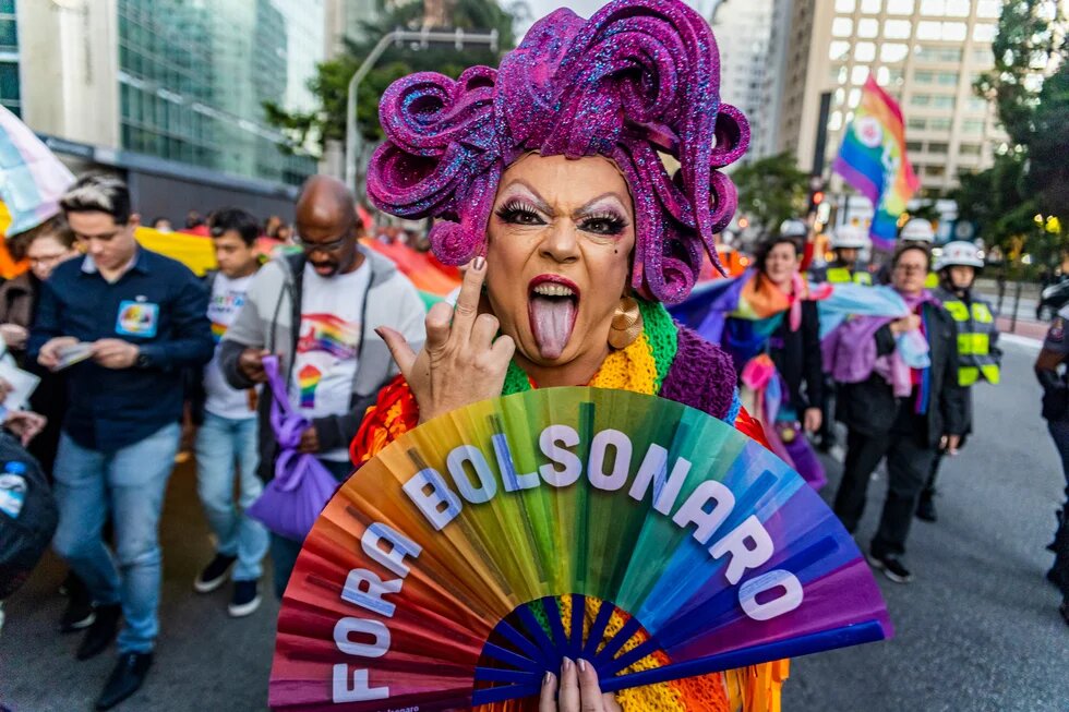 Claude hat geantwortet: Protestfoto von einer Pride-Demonstration in Brasilien (Foto: Mídia Ninja).Protestfoto von einer Pride-Demonstration in Brasilien. Im Vordergrund eine Drag-Person mit pinker Glitzerperücke, dramatischem Make-up und buntem Outfit, die einen Regenbogenfächer mit der Aufschrift „FORA BOLSONARO" hält und die Zunge herausstreckt. Im Hintergrund Demonstrierende mit Regenbogenfahnen und Pride-T-Shirts sowie Polizeibeamt*innen.