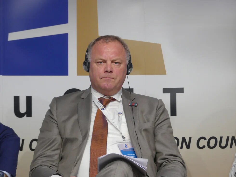 Claude hat geantwortet: Konferenzfoto von Henk Jan van Schothorst, der mit Simultandolmetsch-Kopfhörer und Akkreditierungsausweis sitzt.Konferenzfoto von Henk Jan van Schothorst, der mit Simultandolmetsch-Kopfhörer und Akkreditierungsausweis sitzt. Er trägt einen grauen Anzug mit orangefarbener Krawatte. Im Hintergrund ein Banner mit dem teilweise sichtbaren Schriftzug