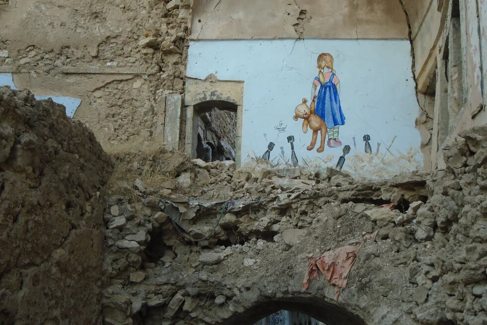 Inmitten von Trümmern und zerstörten Gebäuden ist an einer noch stehenden Wand ein farbiges Wandgemälde zu sehen: ein kleines Mädchen im blauen Kleid, das einen Teddybären hält und mit dem Rücken zur Betrachterin steht, umgeben von stilisierten Bomben oder Raketen am Boden.